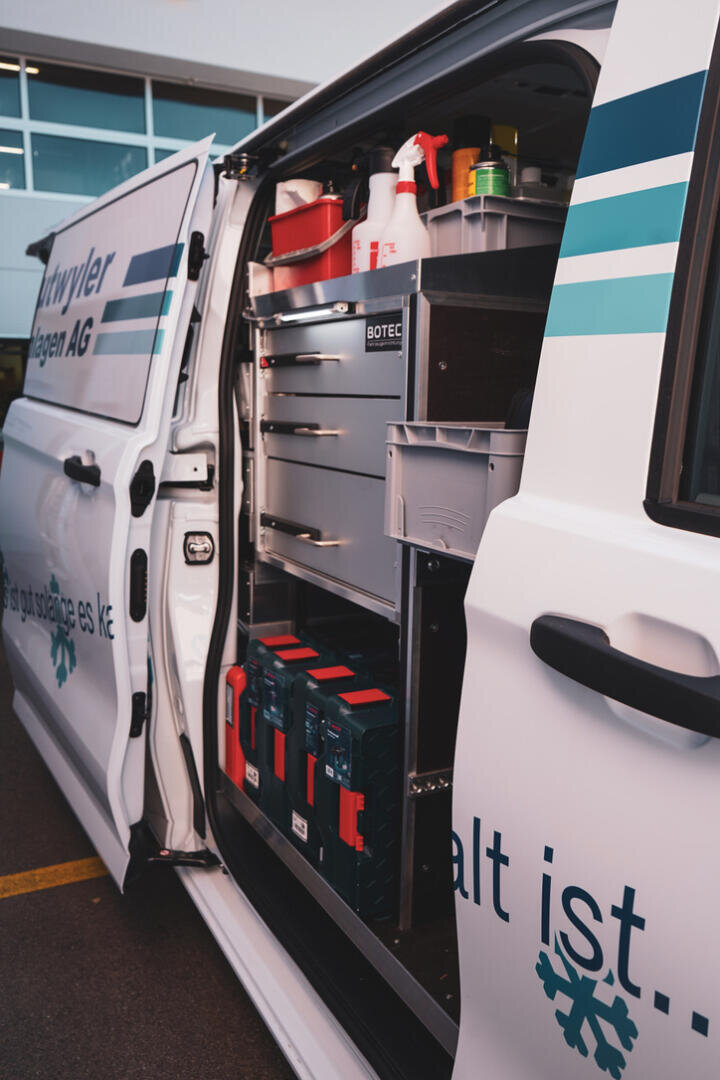 Ein weißer Servicewagen mit geöffneter Seitentür gibt den Blick frei auf geordnete Regale, Schubladen und Aufbewahrungsboxen, die mit Werkzeugen, Reinigungsmitteln und Geräten gefüllt sind und vor einem Gebäude geparkt sind.