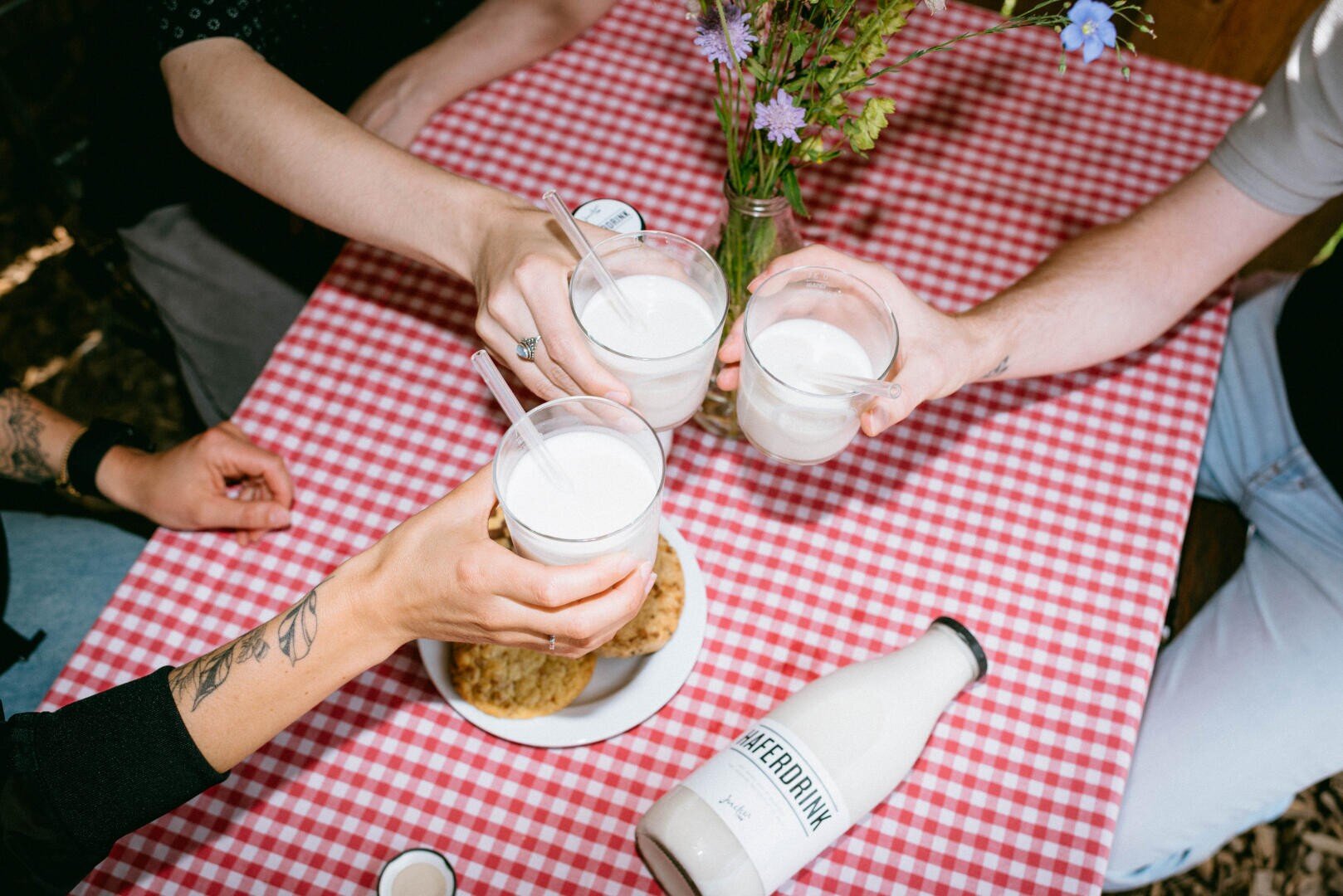 Quatre personnes portent un toast avec des verres d'une boisson blanche sur une nappe à carreaux rouges. Un vase de fleurs, une assiette de biscuits et une bouteille étiquetée #HERDRINK sont posés sur la table.