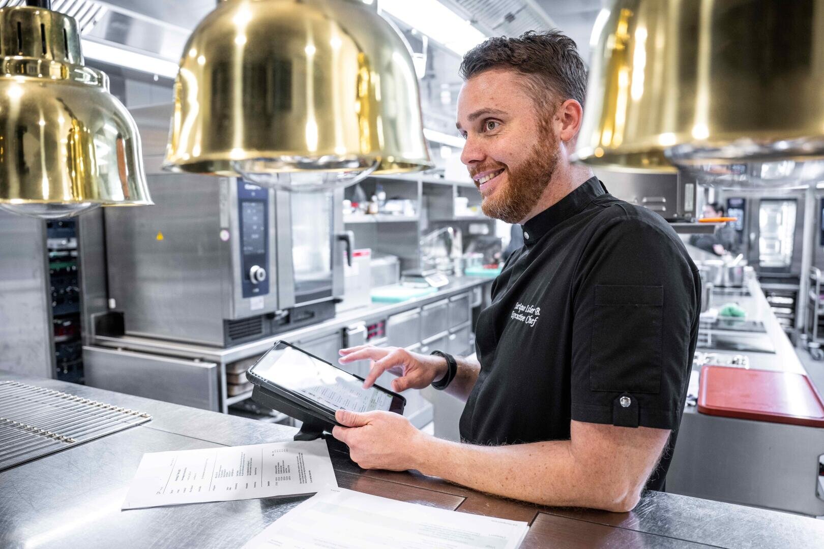 Un chef cuisinier barbu se tient dans une cuisine moderne en acier inoxydable et sourit en utilisant une tablette. Des papiers sont posés sur le comptoir devant lui et des lampes suspendues dorées sont suspendues au-dessus de lui.