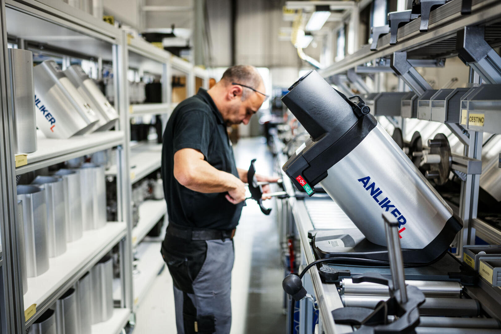 Une personne en tenue de travail monte ou vérifie une grande machine métallique portant l'inscription ANLIKER à un poste de travail dans une usine ou un atelier, entourée d'appareils et de pièces similaires sur des étagères.