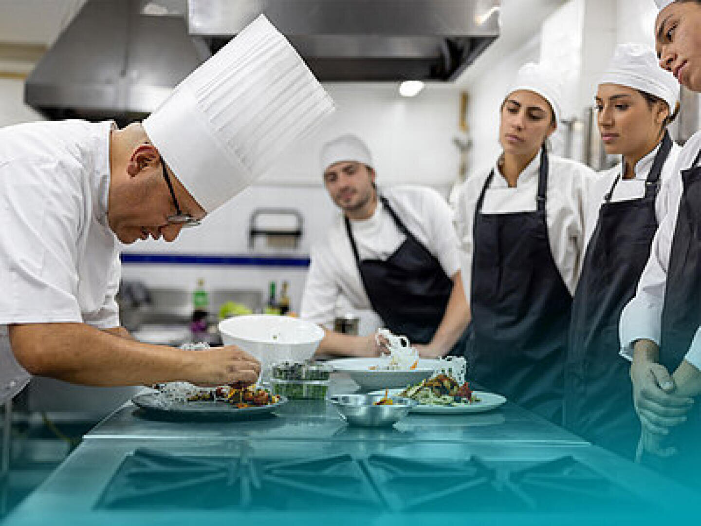 Un chef en toque blanche et en uniforme prépare soigneusement les plats, tandis que trois élèves cuisiniers en tablier et casquette observent attentivement dans une cuisine professionnelle.