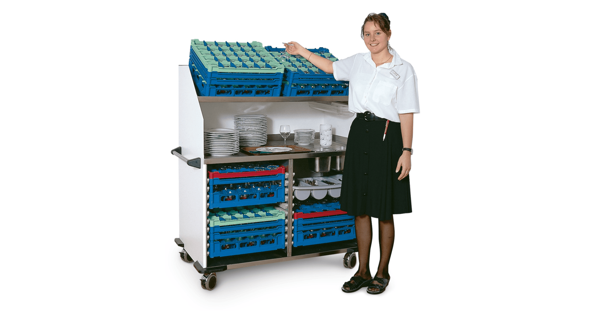 Une femme en chemise blanche et jupe noire se tient à côté d'un chariot mobile rempli d'étagères bleues contenant des assiettes, des tasses et des verres, elle sourit et désigne l'étagère supérieure.