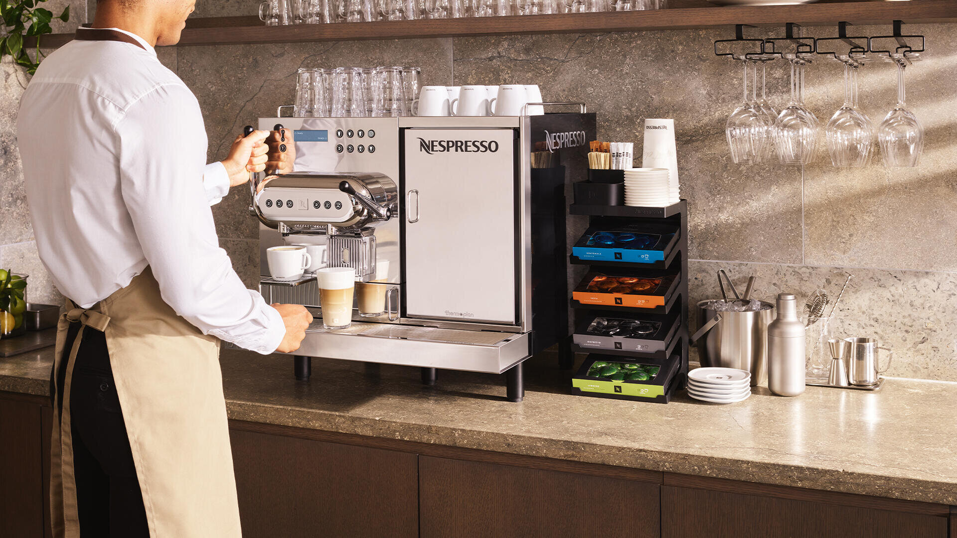 Eine Person in einer Schürze bedient eine Nespresso-Kaffeemaschine in einer modernen Küche und bereitet Getränke zu, wobei Tassen, Tabletts mit Kaffeepads und gestapelte Gläser auf der Arbeitsplatte zu sehen sind.