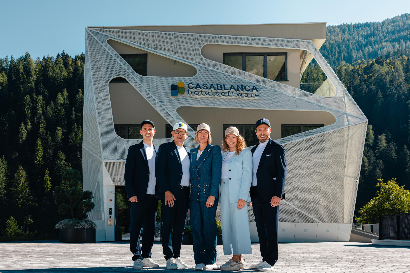 Fünf Personen in Geschäftskleidung und mit Hut stehen lächelnd vor einem modernen weißen Gebäude mit einem Schild mit der Aufschrift CASABLANCA hotelsoftware. Im Hintergrund sind grüne Bäume und Hügel zu sehen.