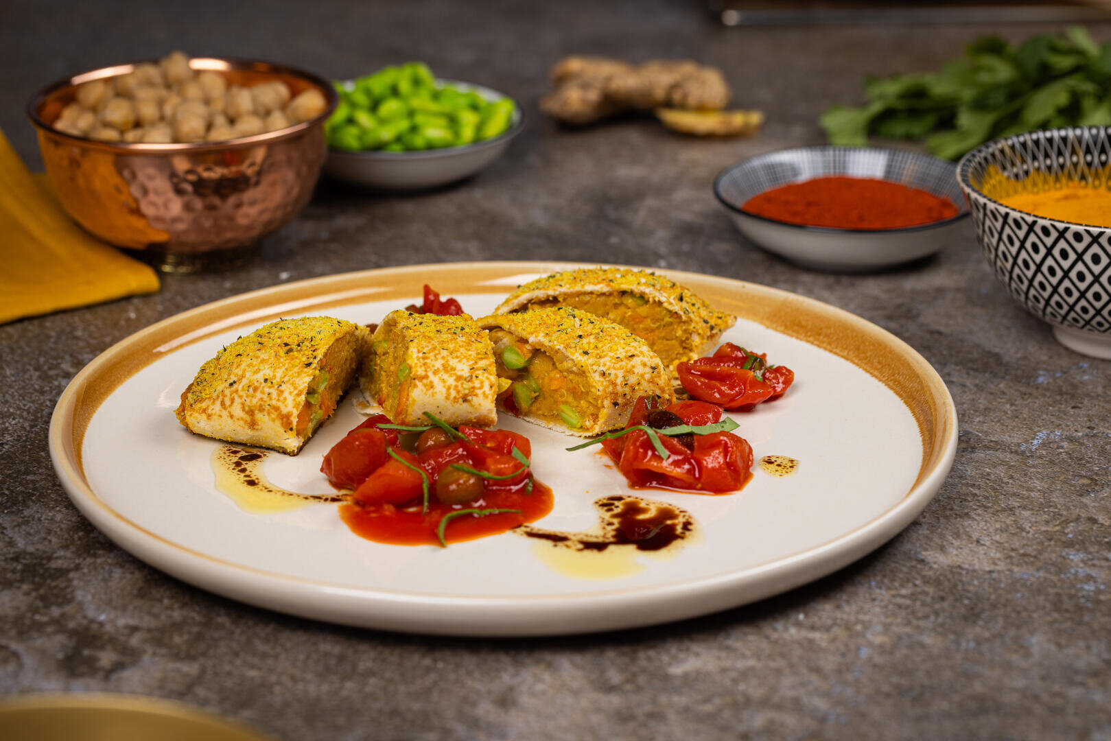 Ein Teller mit gefüllten Teigscheiben, garniert mit gerösteten Kirschtomaten, Soßenspritzern und frischen Kräutern. Im Hintergrund stehen Schalen mit Kichererbsen, Gewürzen und grünem Gemüse auf einer Steinplatte.