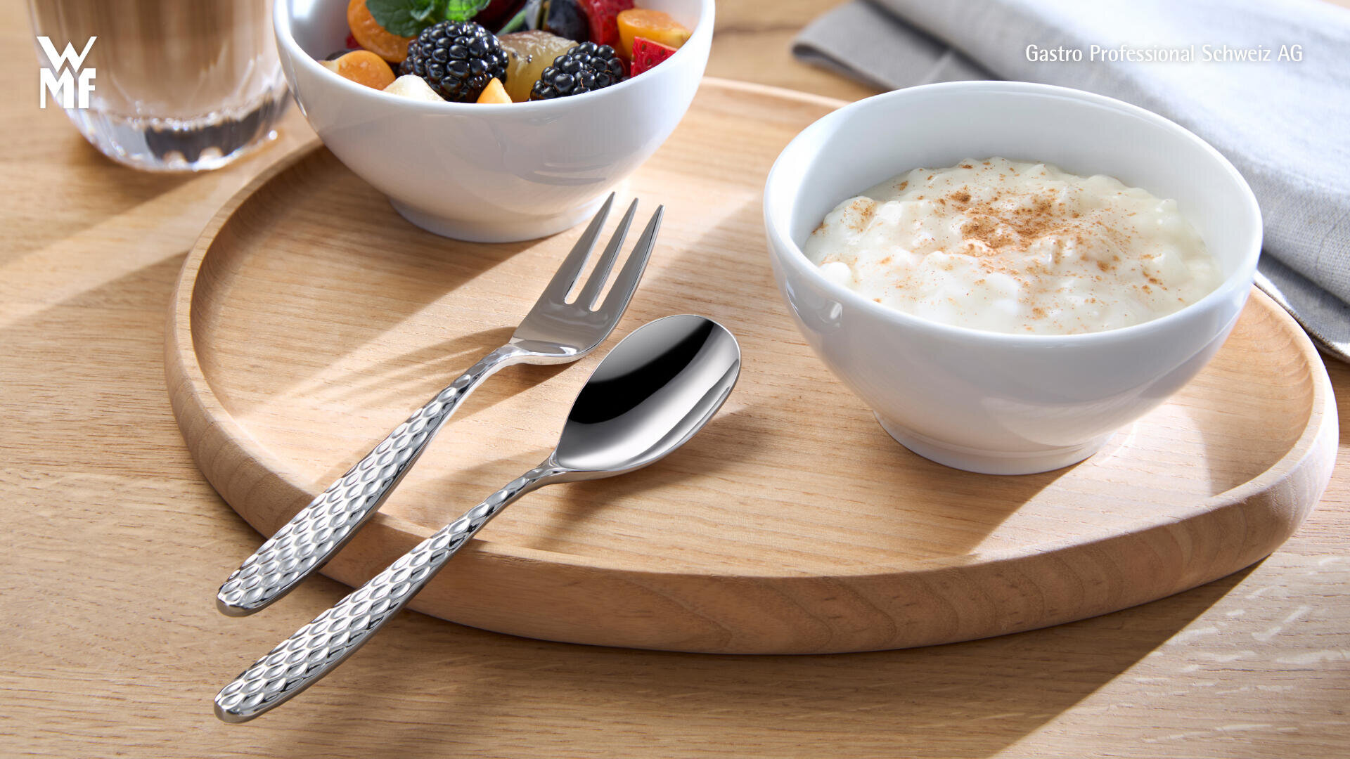 Sur un plateau rond en bois se trouve un bol de riz au lait saupoudré de cannelle, à côté d'un bol de fruits frais mélangés, une fourchette et une cuillère aux manches structurés.