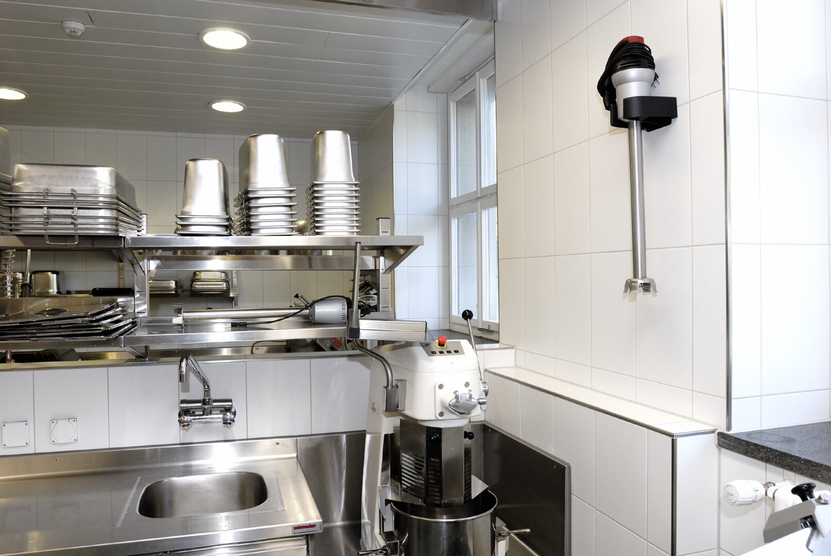 Une cuisine professionnelle épurée avec des appareils électroménagers en acier inoxydable, des récipients métalliques empilés, un grand mixeur industriel et un interrupteur d'arrêt d'urgence mural près d'une fenêtre. Des murs carrelés blancs et des plafonniers sont visibles.