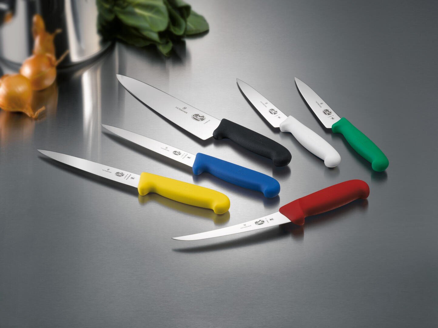Sechs Küchenmesser mit farbenfrohen Griffen (gelb, blau, schwarz, weiß, grün, rot) sind auf einer spiegelnden Oberfläche aus rostfreiem Stahl angeordnet, mit einigen Zwiebeln und Blattgemüse im Hintergrund.