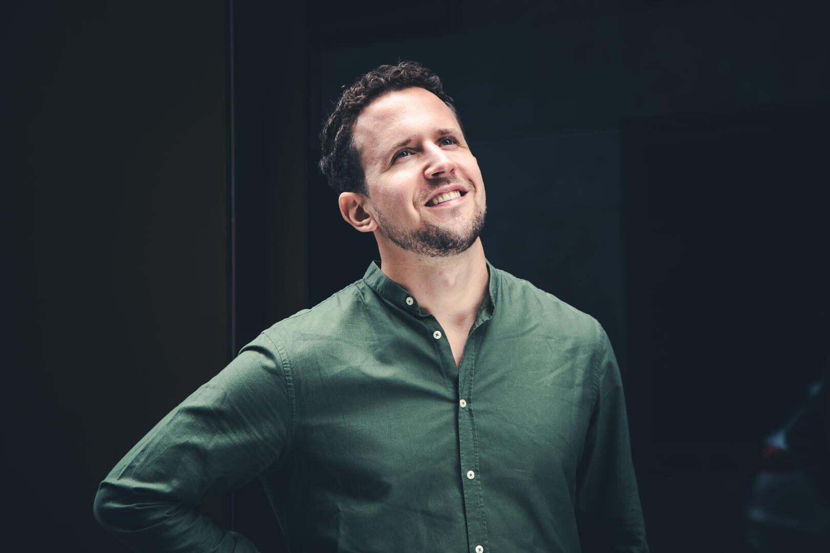 Ein Mann mit kurzen braunen Haaren und gestutztem Bart, der ein grünes Button-up-Hemd trägt, steht lächelnd und mit einer Hand in der Hüfte vor einem dunklen, verschwommenen Hintergrund.