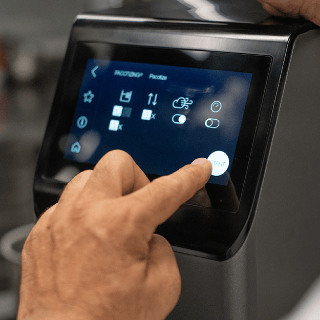 La main d'une personne appuie sur la touche START d'un panneau de commande à écran tactile, qui affiche différentes icônes et réglages, éventuellement pour un appareil de cuisine ou une machine.