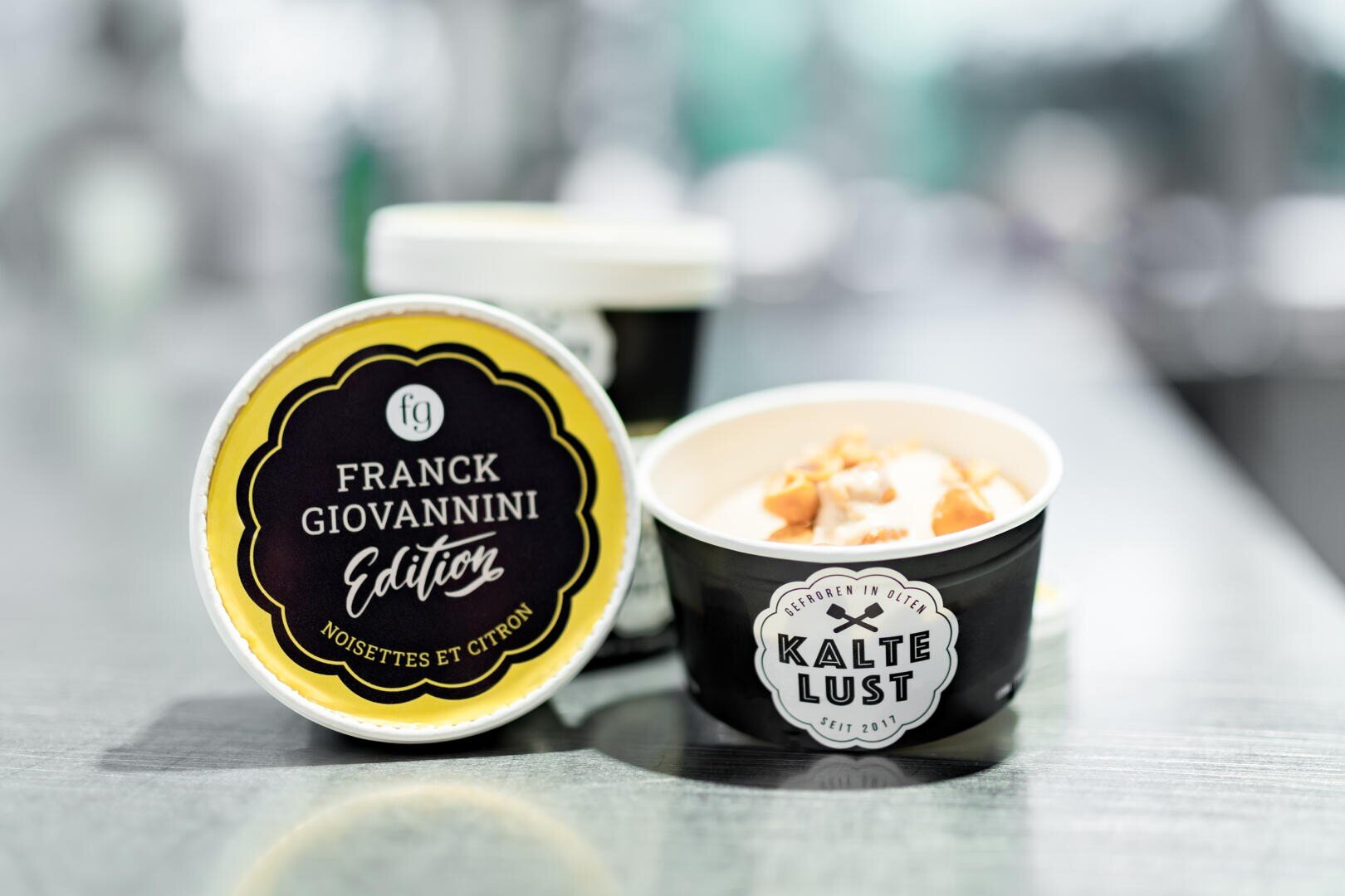 Drei schwarze Eiscremebehälter mit der Aufschrift Franck Giovannini Edition Noisettes et Citron und Kalte Lust sind auf einer reflektierenden Oberfläche ausgestellt, wobei ein Behälter geöffnet ist und Eis mit Topping zeigt.