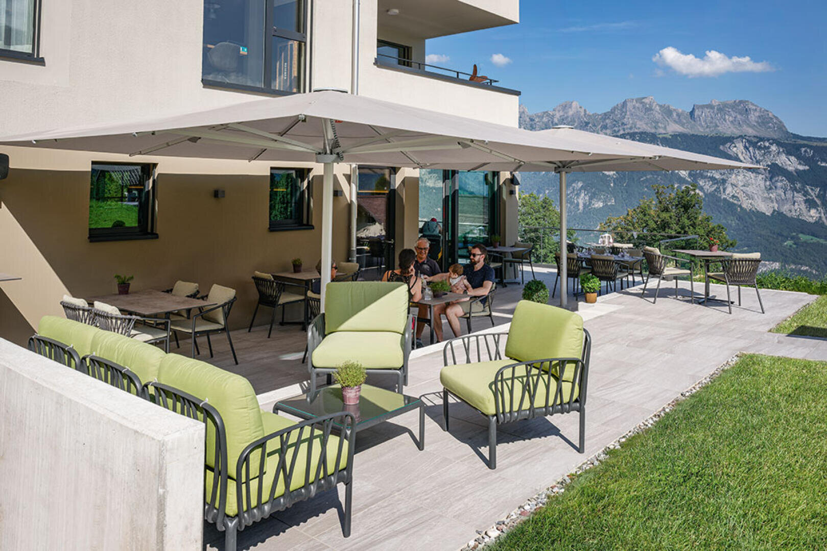 Eine geräumige Außenterrasse mit grünen Sitzkissen, Tischen und einem großen Sonnenschirm. Zwei Personen sitzen lesend zusammen und genießen die Aussicht auf die Berge und das sonnige Wetter. Modernes Gebäude und üppige Landschaft im Hintergrund.