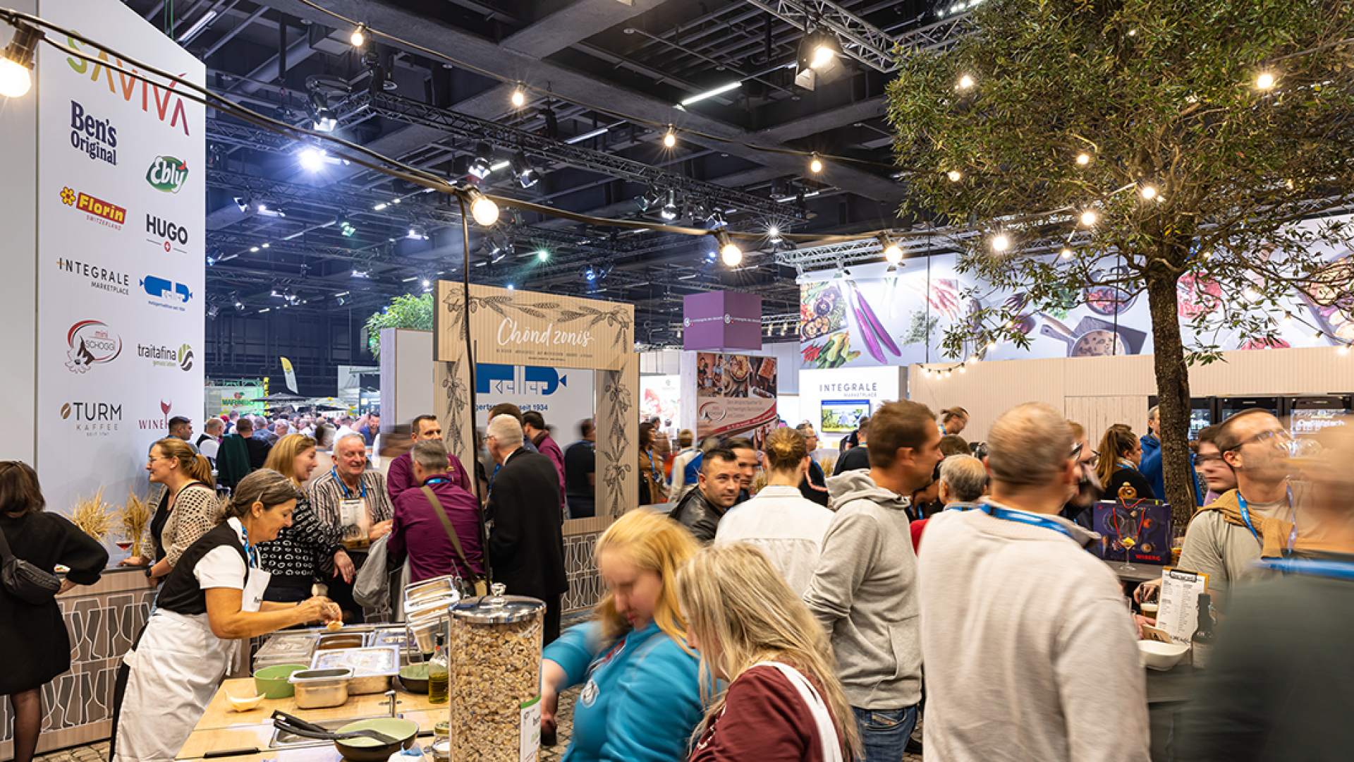 Eine belebte überdachte Lebensmittelausstellung mit Menschenmassen, die an den verschiedenen Ständen vorbeilaufen und Produkte probieren. Große Sponsorenbanner und dekorative Lichter hängen von der Decke und schaffen eine lebhafte Atmosphäre.