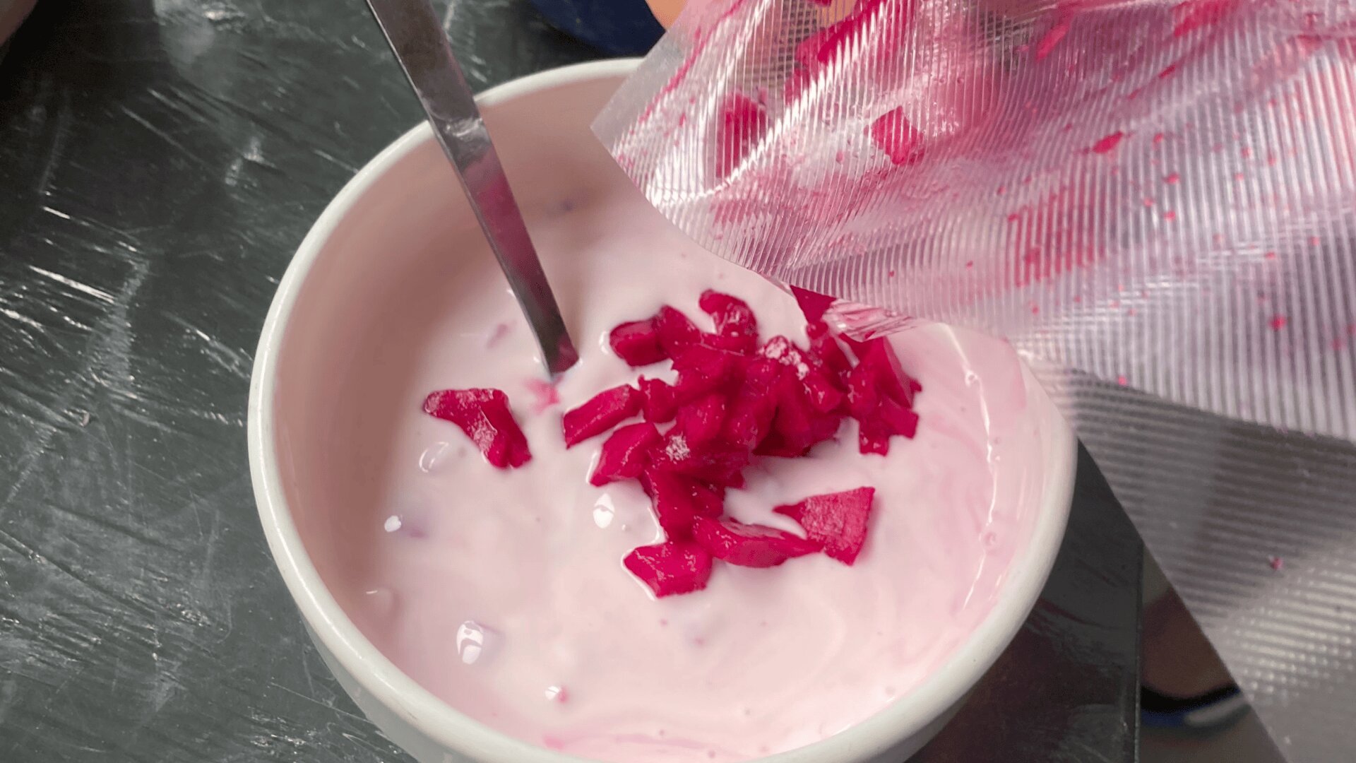 Une main verse des légumes roses hachés d'un sac en plastique dans un bol de yaourt rose clair, avec une cuillère à l'intérieur du bol.