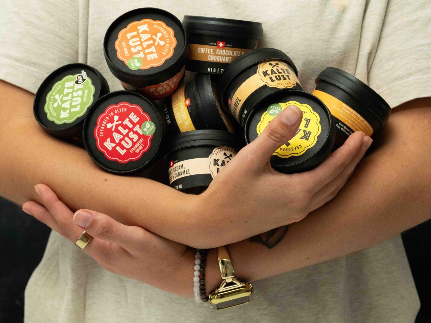 Eine Person in einem hellen Hemd hält mehrere kleine bunte Behälter mit Kaktus-Lust-Eis in ihren Armen, auf denen verschiedene Geschmacksrichtungen zu sehen sind. Auch die Uhr und das Armband der Person sind zu sehen.