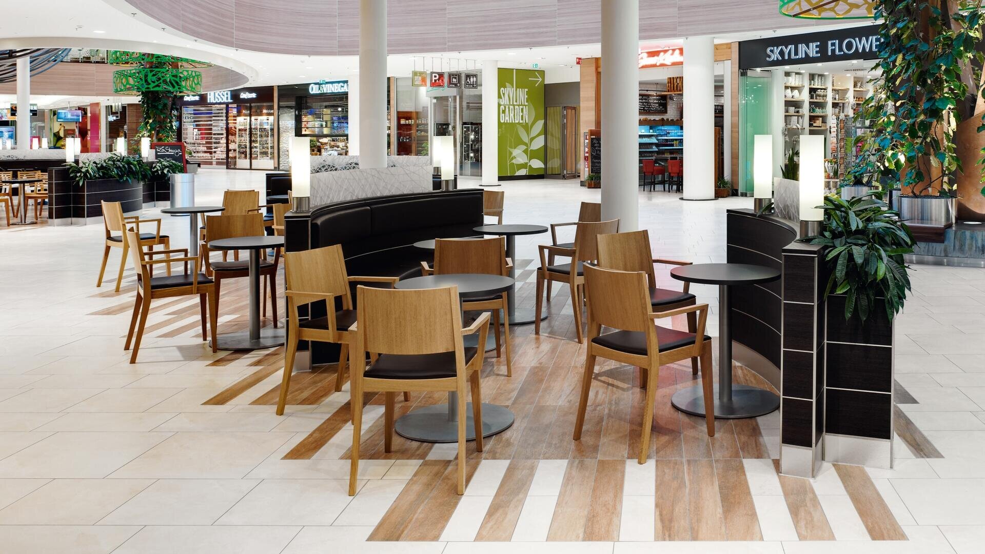 Une aire de restauration intérieure moderne avec des tables et des chaises en bois vides, un sol en carrelage clair et des devantures de magasins en arrière-plan, dont un fleuriste et un café. Des plantes vertes ajoutent une touche de nature à l'espace.