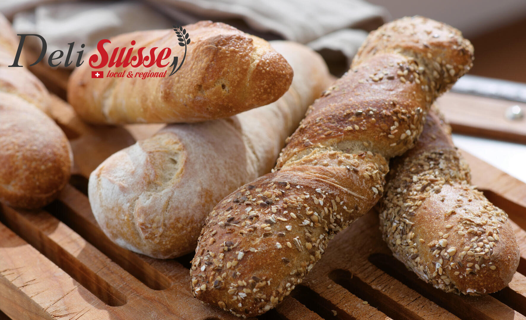Ein Holzbrett mit drei rustikalen Baguettes, zwei davon mit Körnern belegt, nebeneinander. In der oberen linken Ecke ist das Logo "Deli Suisse local & regional" zu sehen.