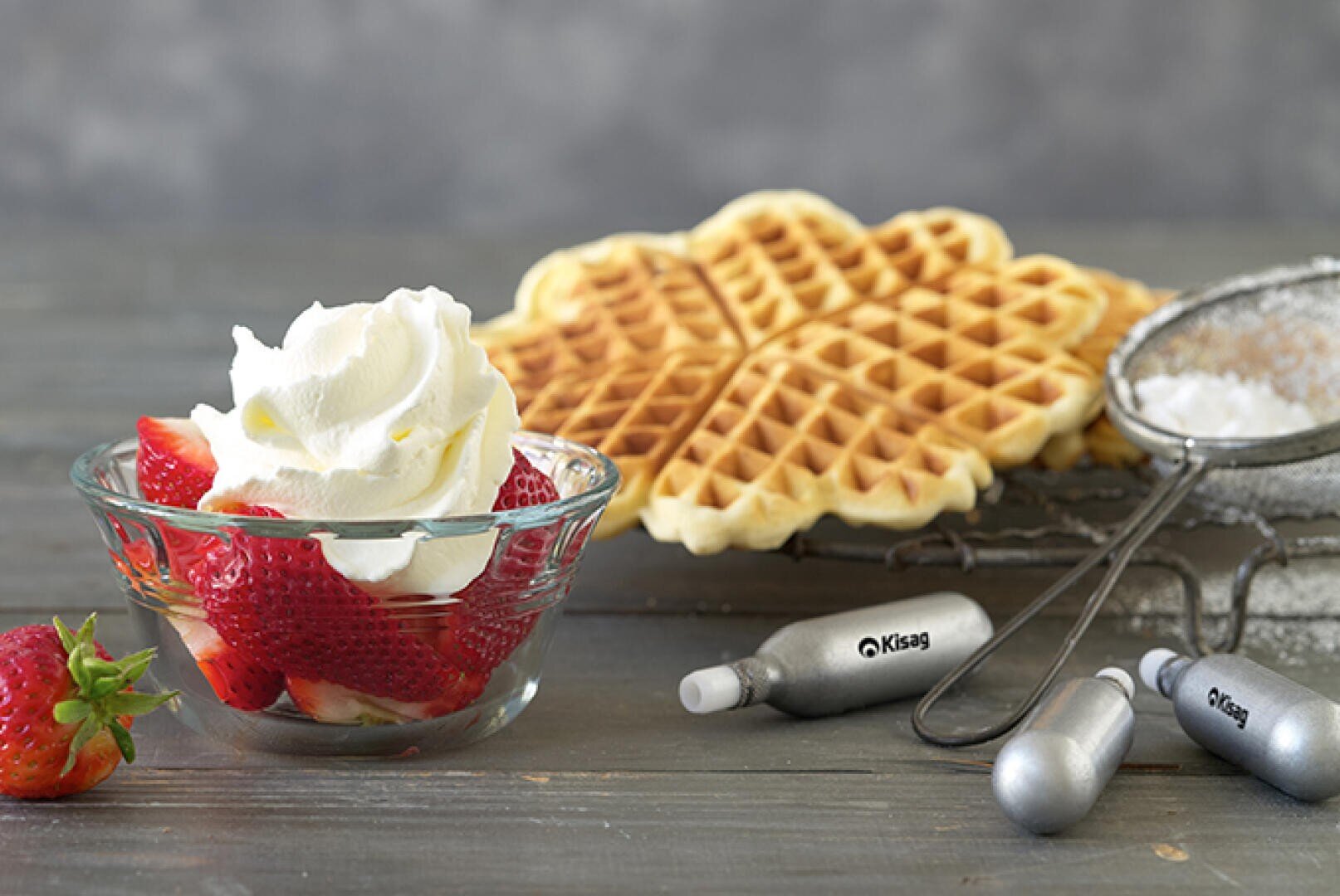 Eine Glasschale gefüllt mit geschnittenen Erdbeeren und garniert mit Schlagsahne steht neben herzförmigen Waffeln, einem Metallsieb mit Puderzucker und Sahnekapseln auf einer grauen Oberfläche.