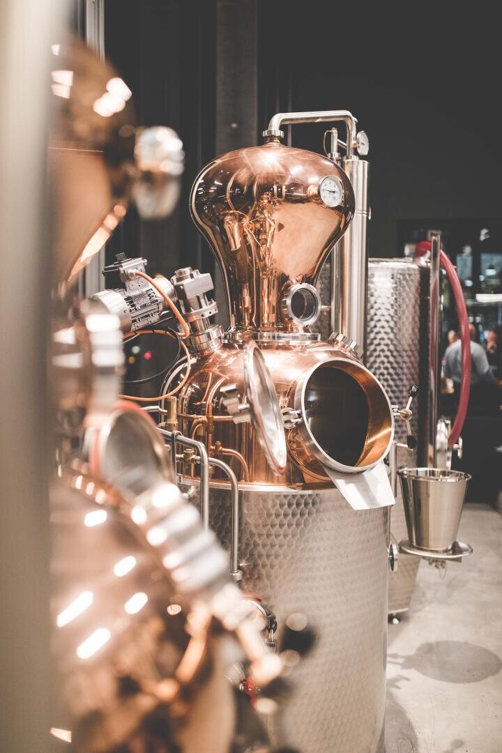 Ein glänzender Destillierapparat aus Kupfer und Edelstahl mit Rohren, Messinstrumenten und einem runden Glasfenster in einer industriellen Umgebung mit sanfter Beleuchtung.