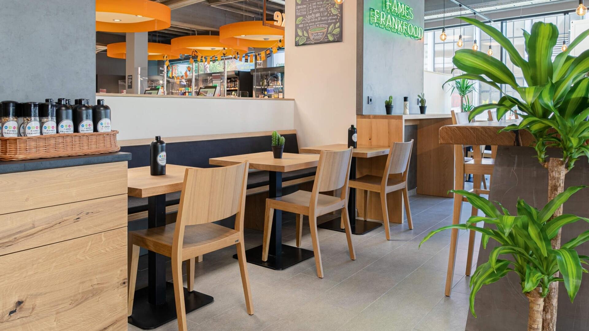 Café moderne avec des tables et des chaises en bois, des plantes en pot et un intérieur clair et propre. Des lumières rondes orange sont suspendues au plafond et une enseigne verte au néon est fixée au mur. De grandes fenêtres laissent entrer la lumière naturelle.