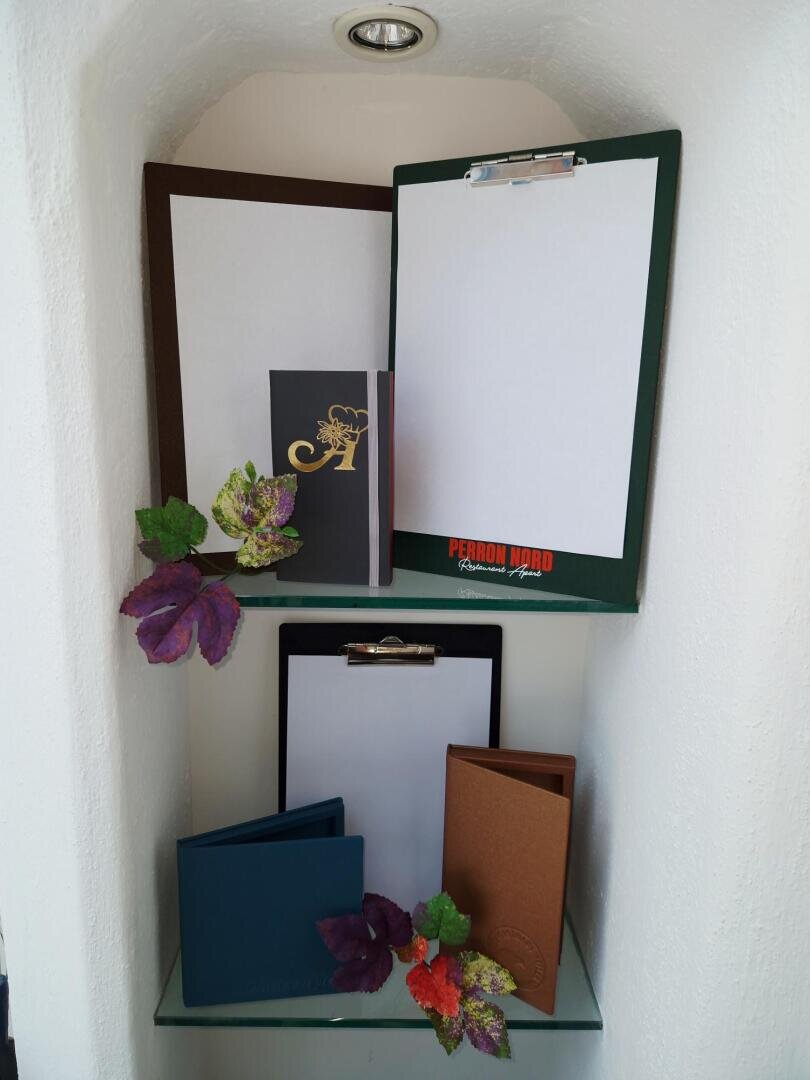 In einem Regal stehen Klemmbretter mit leeren Blättern und geschlossenen Speisekarten, dekoriert mit künstlichen Blättern und Blumen vor einer weißen Wand.