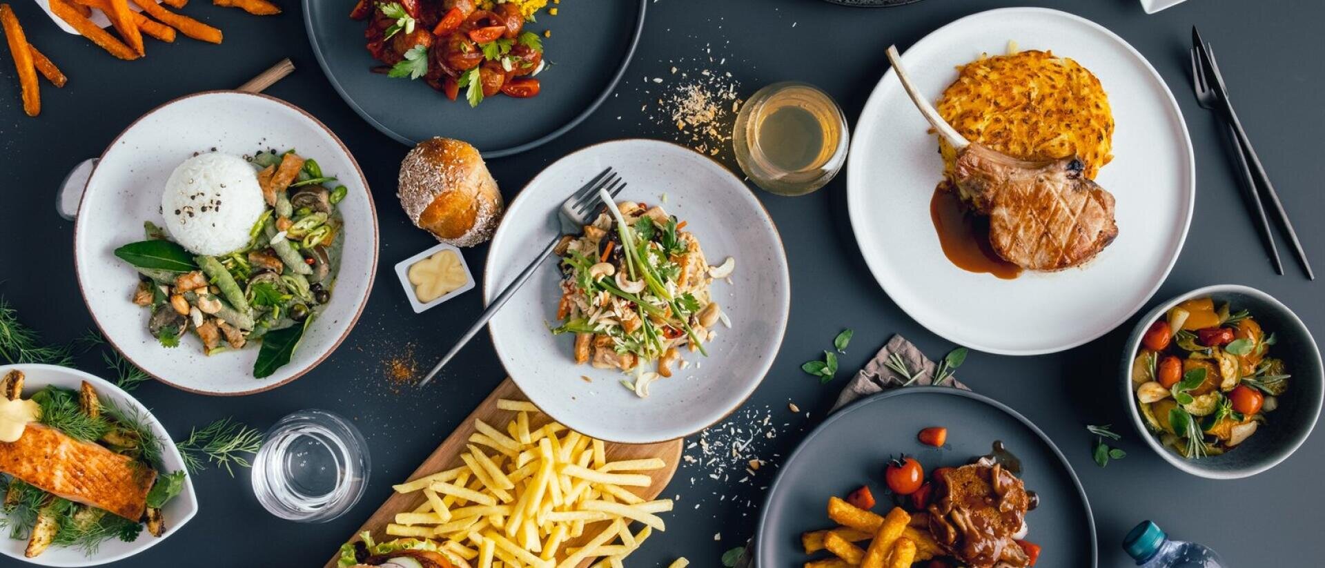 Eine Vielzahl von Gourmetgerichten auf einem dunklen Tisch, darunter Pommes frites, Salat, gebratenes Fleisch, Reis, Gemüse, Brot, ein Gebäckstück und mehrere kleine Schalen mit bunten Beilagen. Auch ein Glas mit Getränken ist zu sehen.