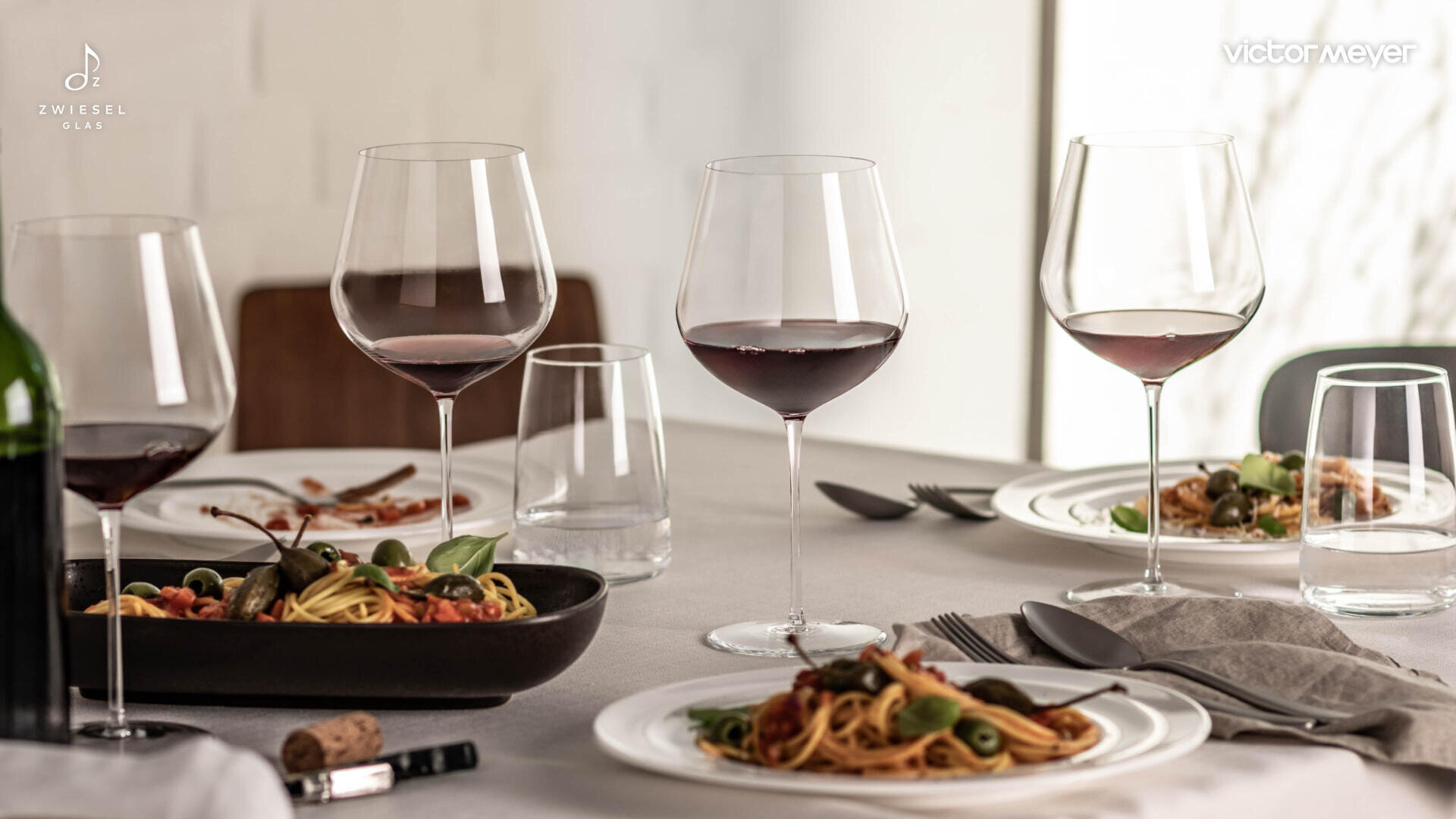 Ein Esstisch mit Pastatellern, drei Rotweingläsern, einem Wasserglas, Besteck und einem Weinkorken, der eine elegante Mahlzeit darstellt.