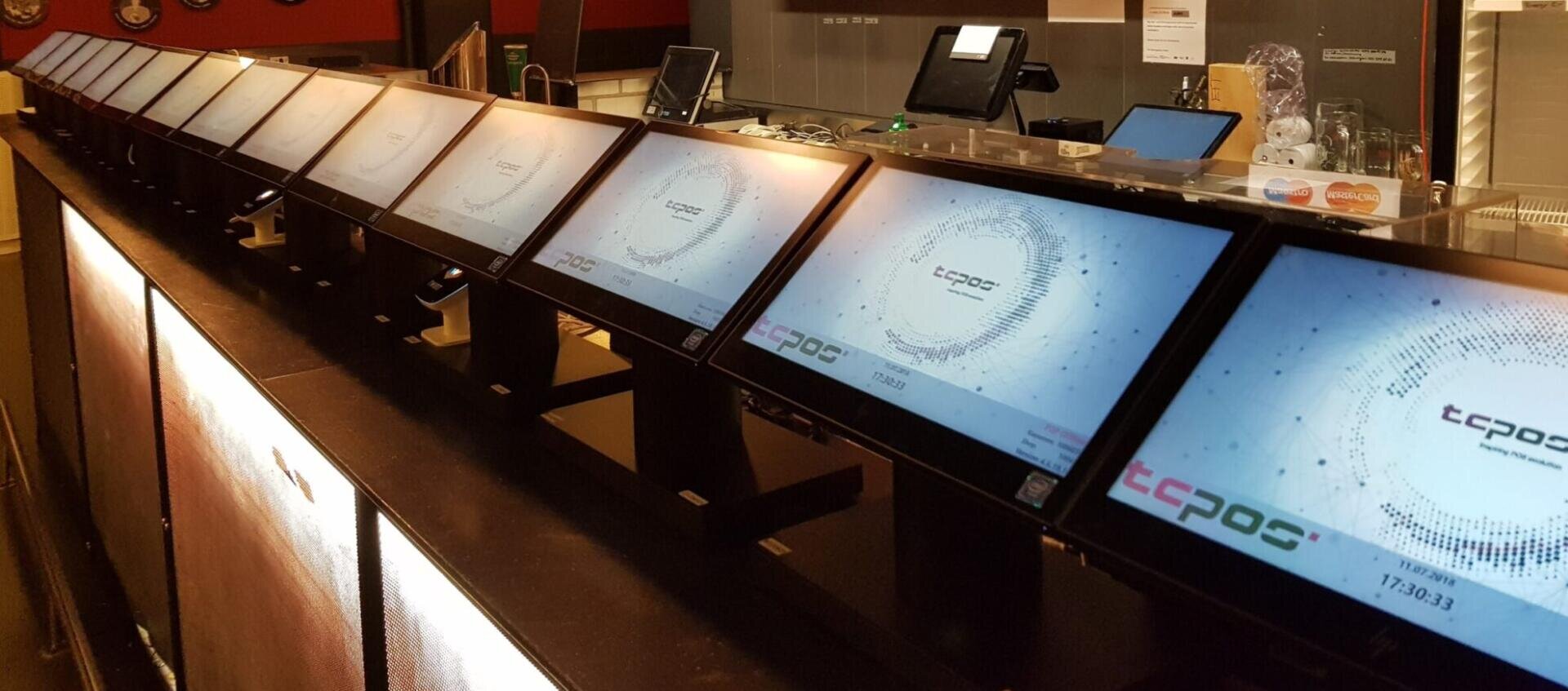Eine Reihe von POS-Terminals (Point-of-Sale) mit Touchscreen auf einer Theke in einem modernen Einzelhandels- oder Restaurantbetrieb, die alle den TCPOS-Startbildschirm anzeigen.