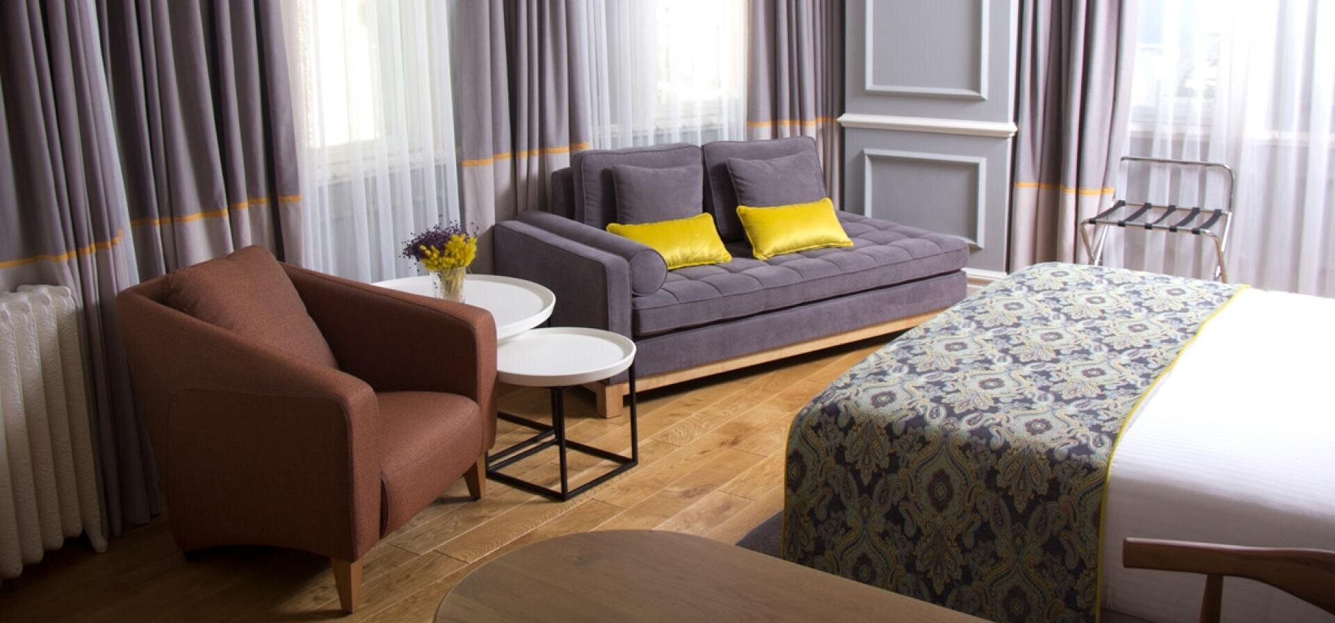 Ein modernes Hotelzimmer mit einer gemusterten Tagesdecke, einem grauen Sofa mit gelben Kissen, einem braunen Sessel, runden Beistelltischen und großen Fenstern mit durchsichtigen Vorhängen, die natürliches Licht hereinlassen.