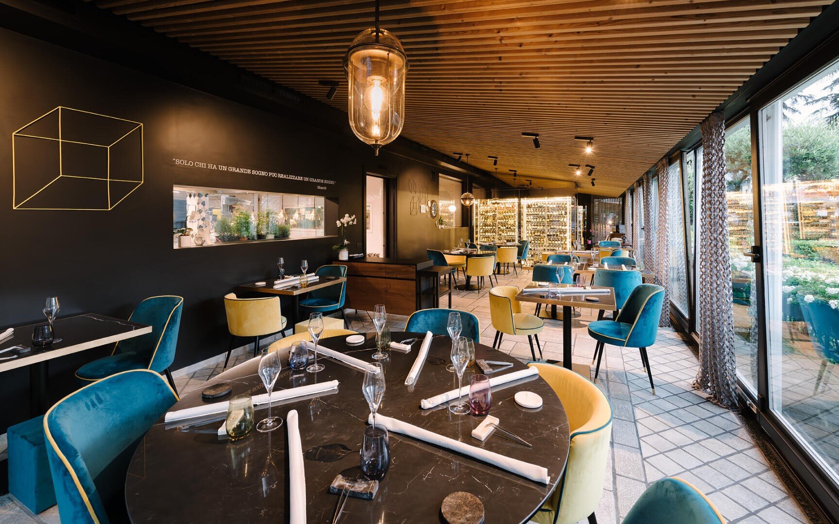 Un intérieur de restaurant moderne avec des tables noires, des chaises bleues et jaunes, de grandes fenêtres laissant entrer la lumière naturelle, et des œuvres d'art murales représentant un cube géométrique et un présentoir de bouteilles de vin en verre.