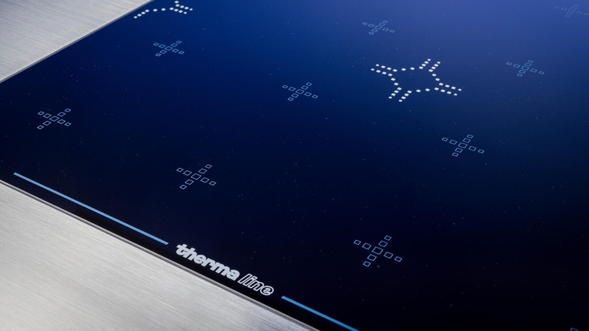 Nahaufnahme eines modernen Induktionskochfelds aus schwarzem Glas mit beleuchteten kreuzförmigen Brenneranzeigen und dem therma line-Logo an der Unterkante.
