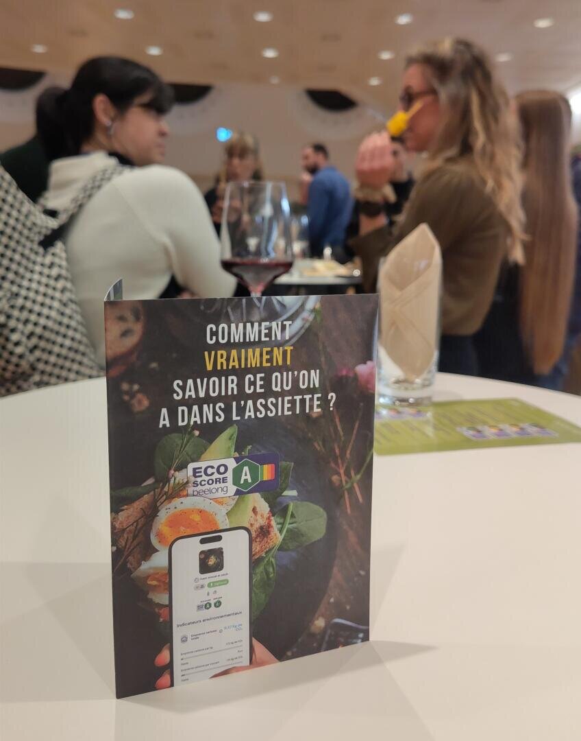 Un panneau de table en français sur la compréhension du contenu de l'assiette, avec une étiquette Eco Score et un téléphone, avec des gens en train de socialiser et un verre de vin en arrière-plan flou.