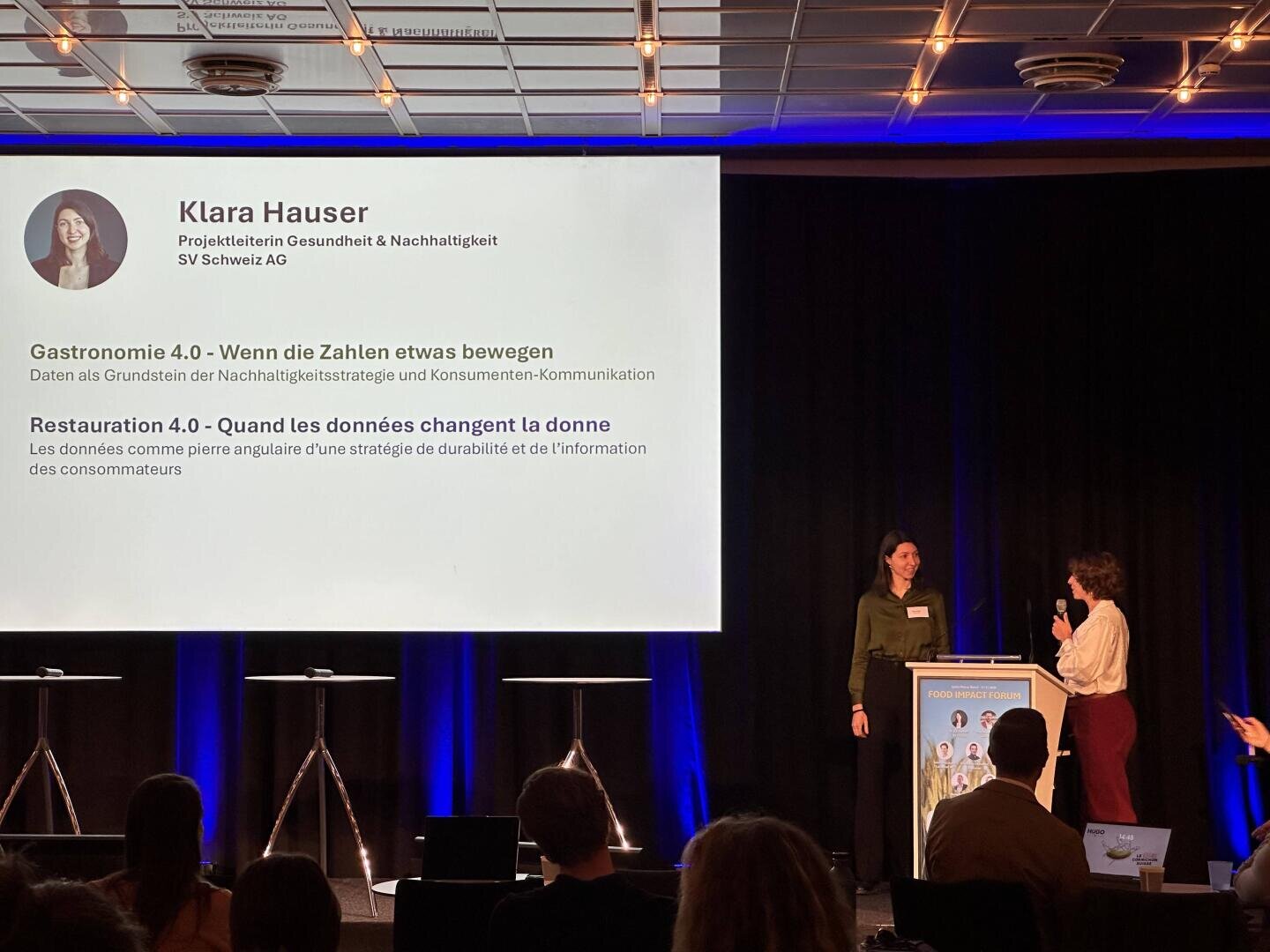 Zwei Frauen stehen auf der Bühne einer Konferenz. Die eine ist Moderatorin, während die andere, Klara Hauser, auf einem Bildschirm hinter ihnen präsentiert wird. Die Folie zeigt ihr Foto, ihren Titel und die Themen ihres Vortrags, die sich auf Nachhaltigkeit und Daten in der Gastronomie beziehen.