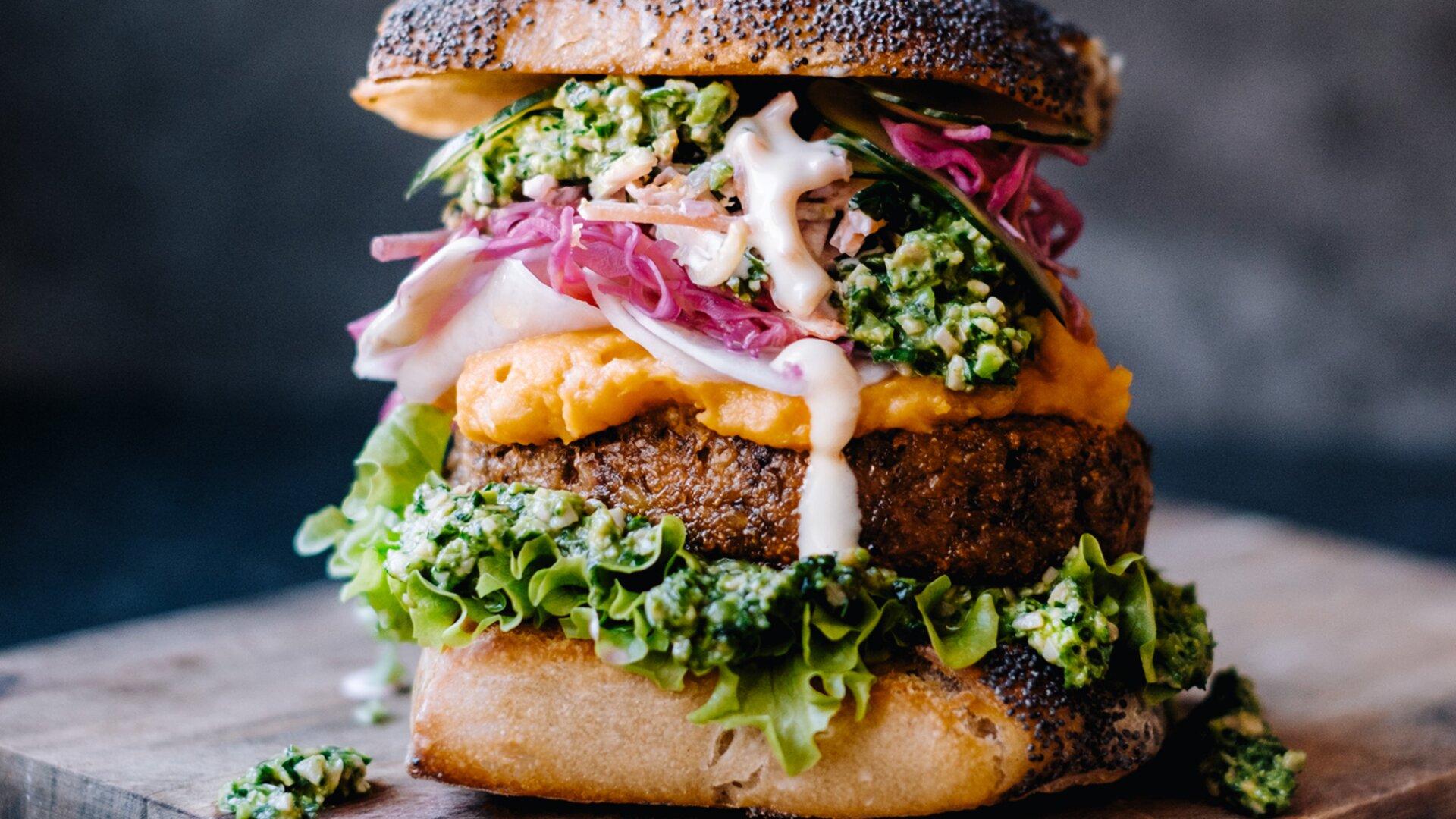 Ein Gourmet-Veggie-Burger mit Salat, Patty, eingelegten Zwiebeln, grünem Pesto und Süßkartoffelpüree in einem knusprigen Brötchen auf einem Holzbrett vor einem dunklen Hintergrund.