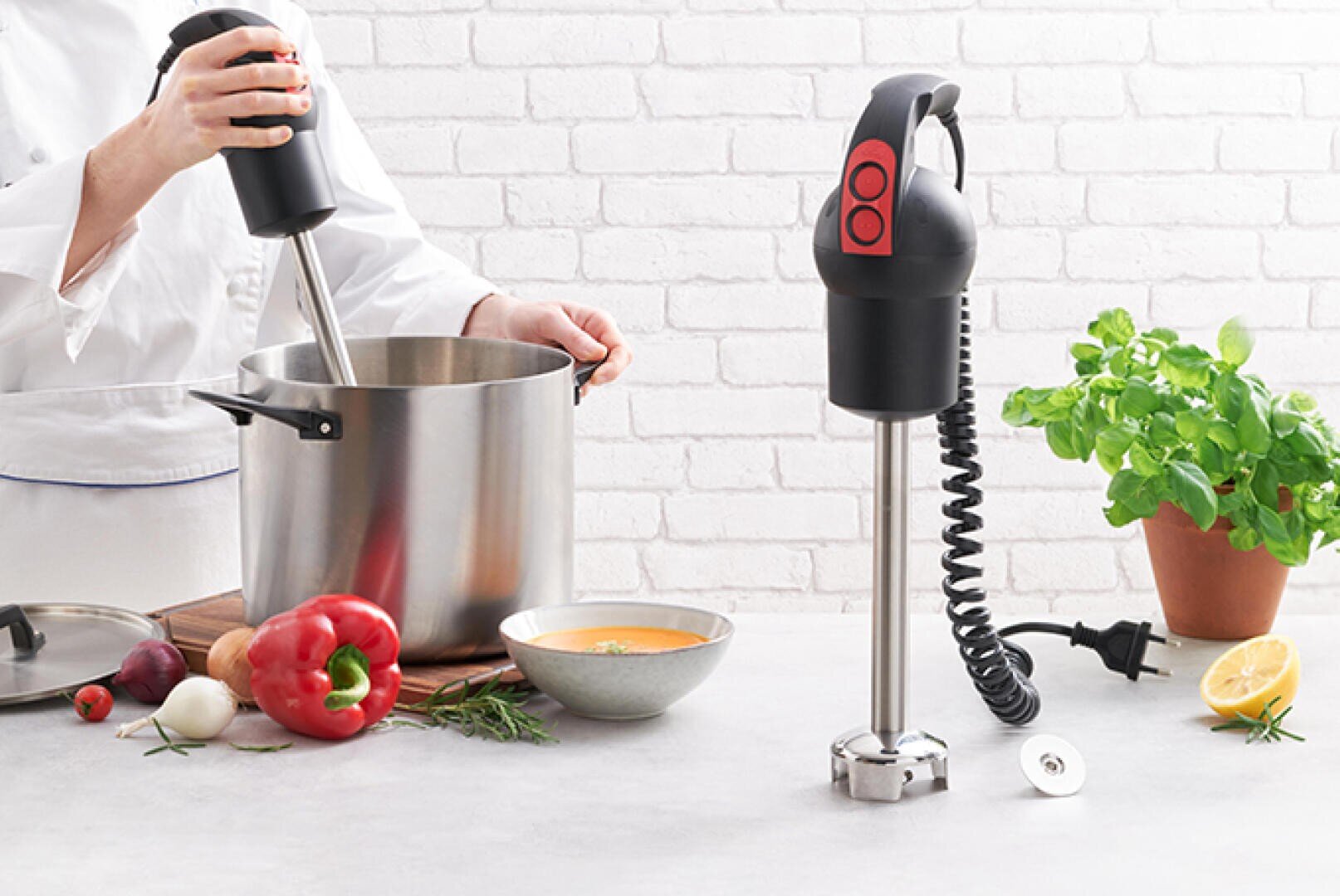 Une personne utilise un mixeur plongeant dans une grande casserole posée sur un plan de travail, avec des légumes, des herbes, un bol de soupe, un plant de basilic en pot et un citron. Un autre mixeur plongeant est placé à la verticale à proximité.