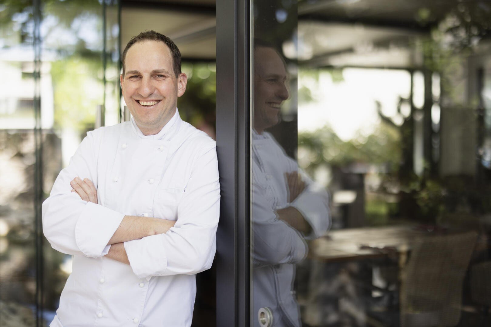 Un cuisinier souriant, vêtu d'un uniforme blanc, se tient debout, les bras croisés, appuyé contre une porte vitrée. Son reflet est visible dans la vitre et, en arrière-plan, on aperçoit un intérieur de restaurant flou.