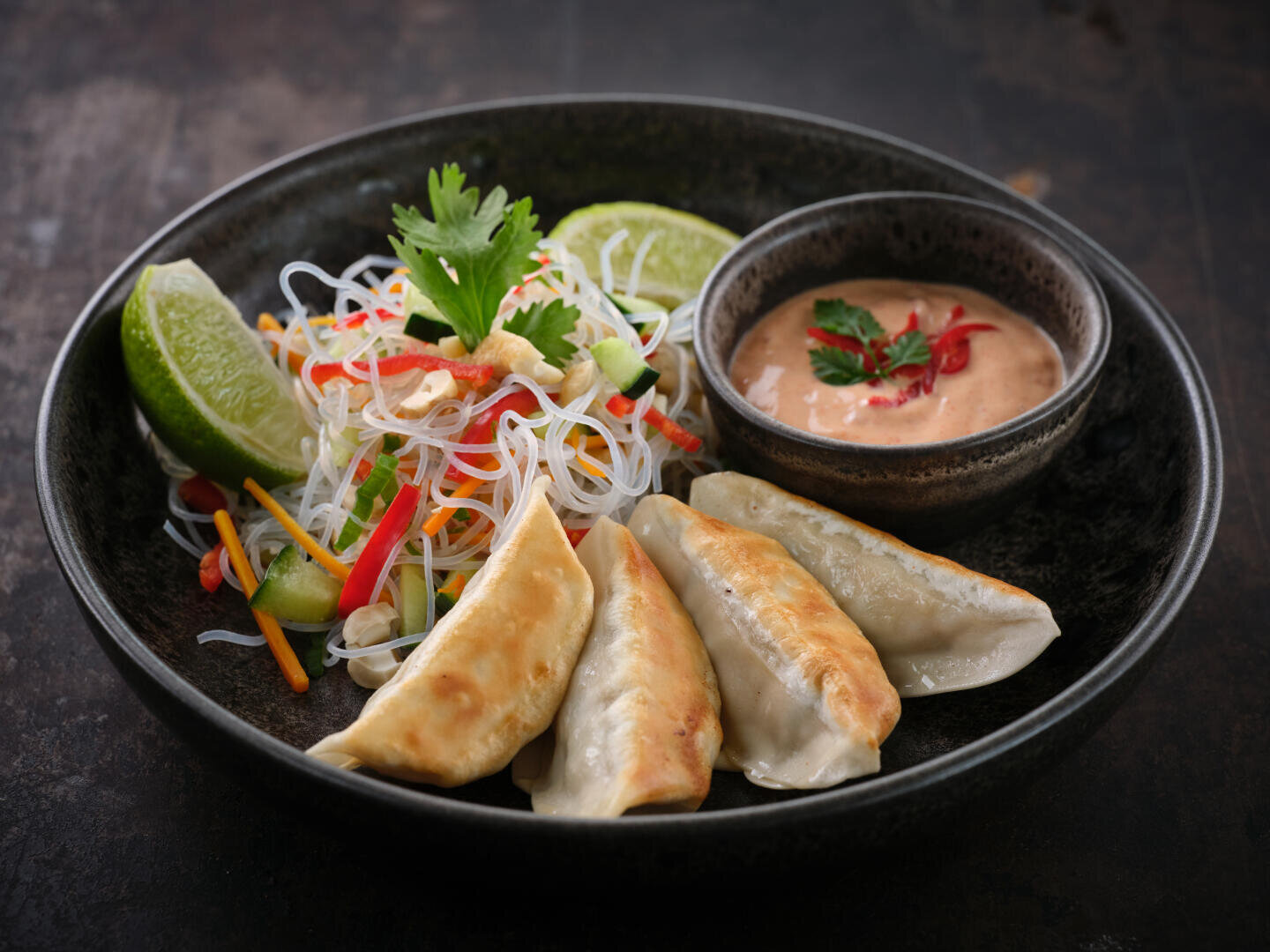 Ein schwarzer Teller mit vier goldgelben, in der Pfanne gebratenen Teigtaschen, ein Fadennudelsalat mit Gemüse und Kräutern, Limettenspalten und eine kleine Schale mit cremiger Dip-Sauce, garniert mit Koriander und rotem Chili.