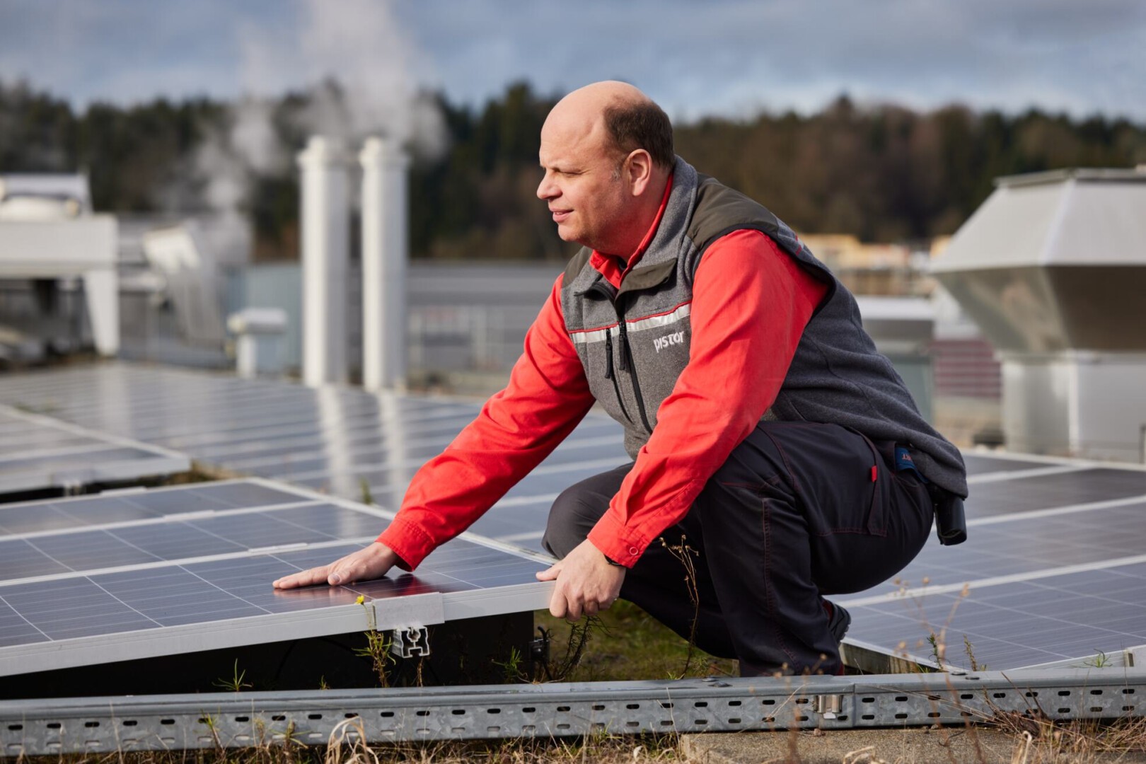 Ein Mann in einem roten Hemd und einer grauen Weste hockt auf einem Dach und inspiziert oder justiert mit einer Hand Solarmodule, im Hintergrund sind Industriegebäude und Bäume zu sehen.