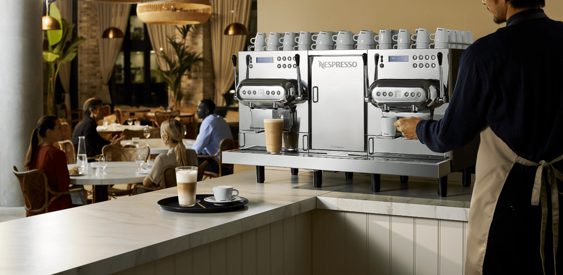 Ein Barista bereitet Kaffee mit einer Nespresso-Maschine in einem Café zu, während die Getränke auf dem Tresen stehen und sich die Leute an den Tischen im Hintergrund unterhalten.