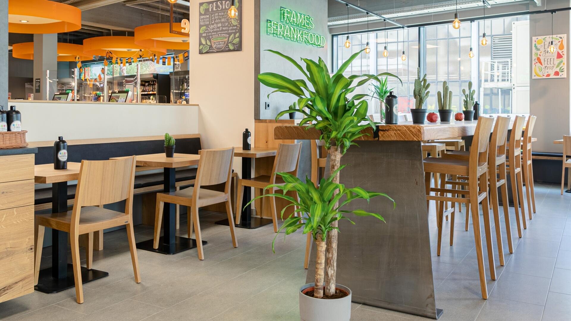 Modernes Restaurant mit Holztischen und -stühlen, hoher Bar, Topfpflanzen, leuchtend orangefarbenen Deckenakzenten und großen Fenstern, die natürliches Licht hereinlassen. Auf dem Neonschild an der Rückwand steht "FRANKS FRANKFURT".