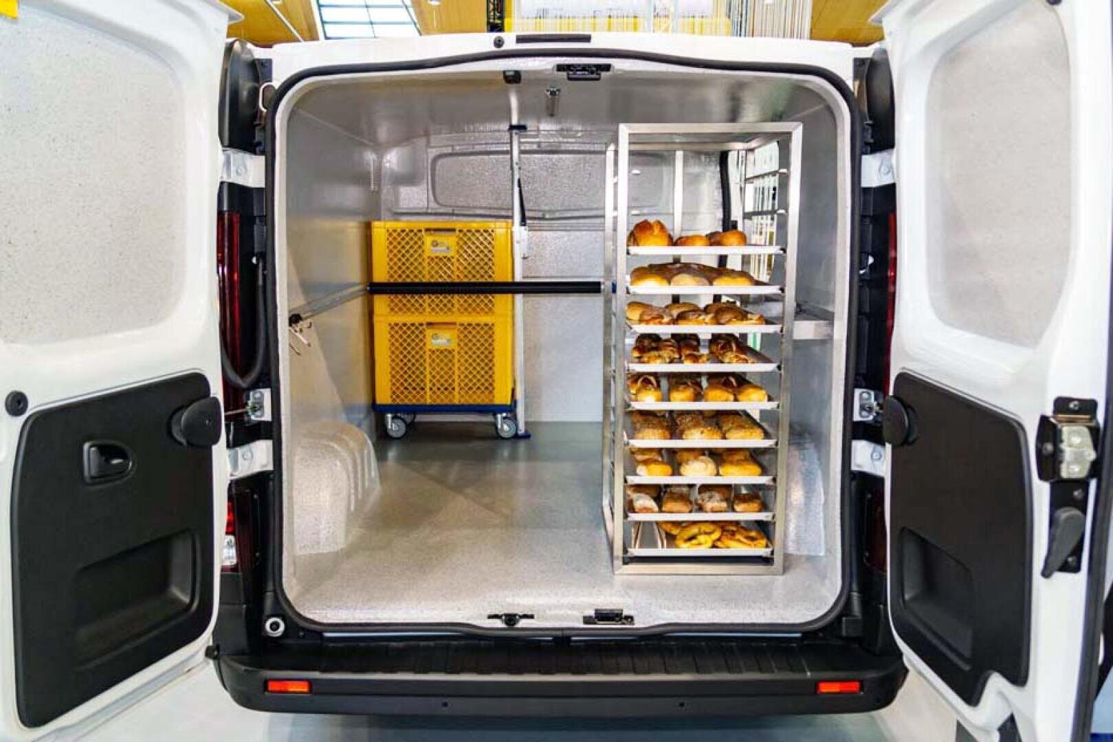 L'image montre les portes arrière ouvertes d'une camionnette réfrigérée. A l'intérieur se trouvent deux caisses jaunes et un châssis métallique, remplis de produits de boulangerie ou de pain et rangés pour le transport.