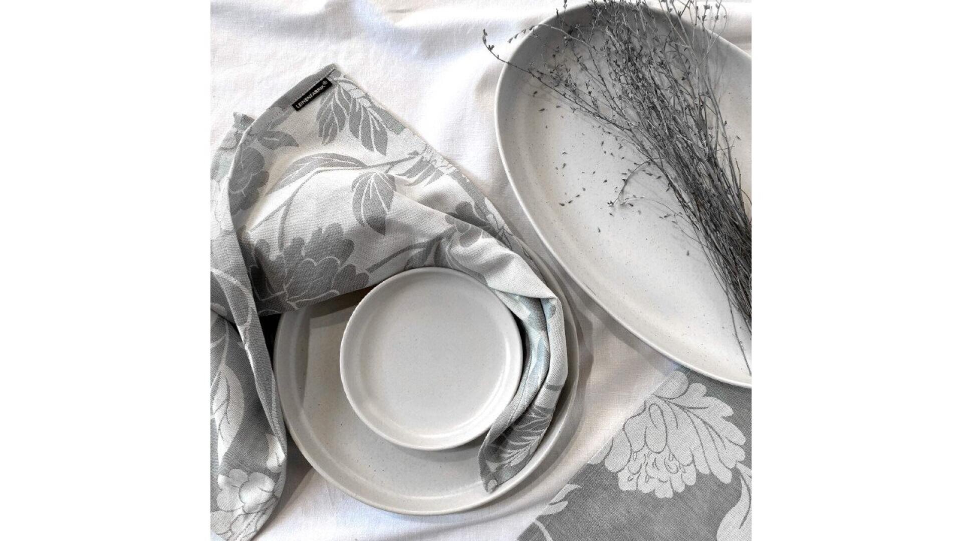 Un ensemble d'assiettes minimalistes en céramique blanche sur un tissu blanc à motifs floraux, avec des brindilles séchées et une serviette de table grise et blanche à motifs floraux élégamment disposée autour des assiettes.