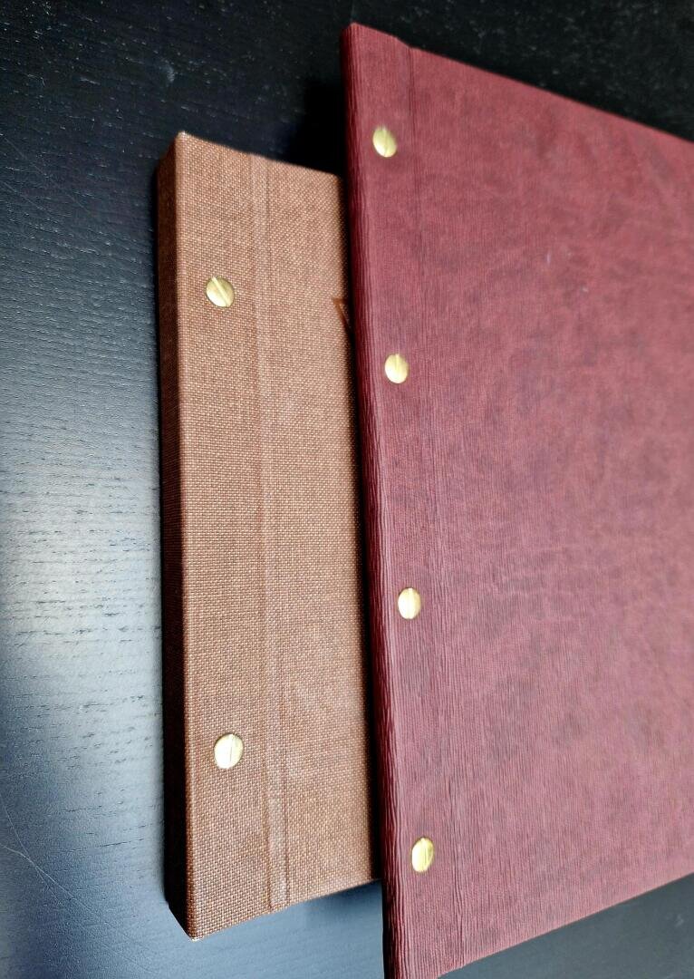 Zwei gebundene Bücher mit braunem und rotem strukturiertem Einband, beide mit drei goldfarbenen Verschlüssen versehen, sind auf einer dunklen Fläche gestapelt. Das rote Buch verdeckt teilweise das braune Buch.