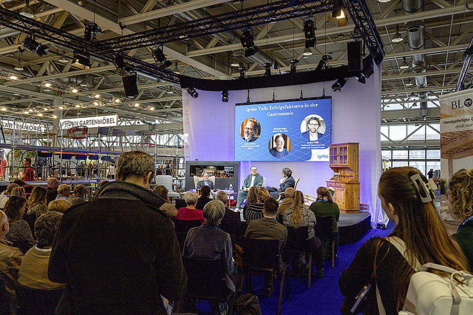 Ein sitzendes Publikum hört einer Podiumsdiskussion auf einer Bühne in einer Kongresshalle zu. Auf der Bühne sind drei Redner zu sehen, deren Bilder und Namen auf einem großen Bildschirm hinter ihnen angezeigt werden. Im Hintergrund sind Messestände zu sehen.