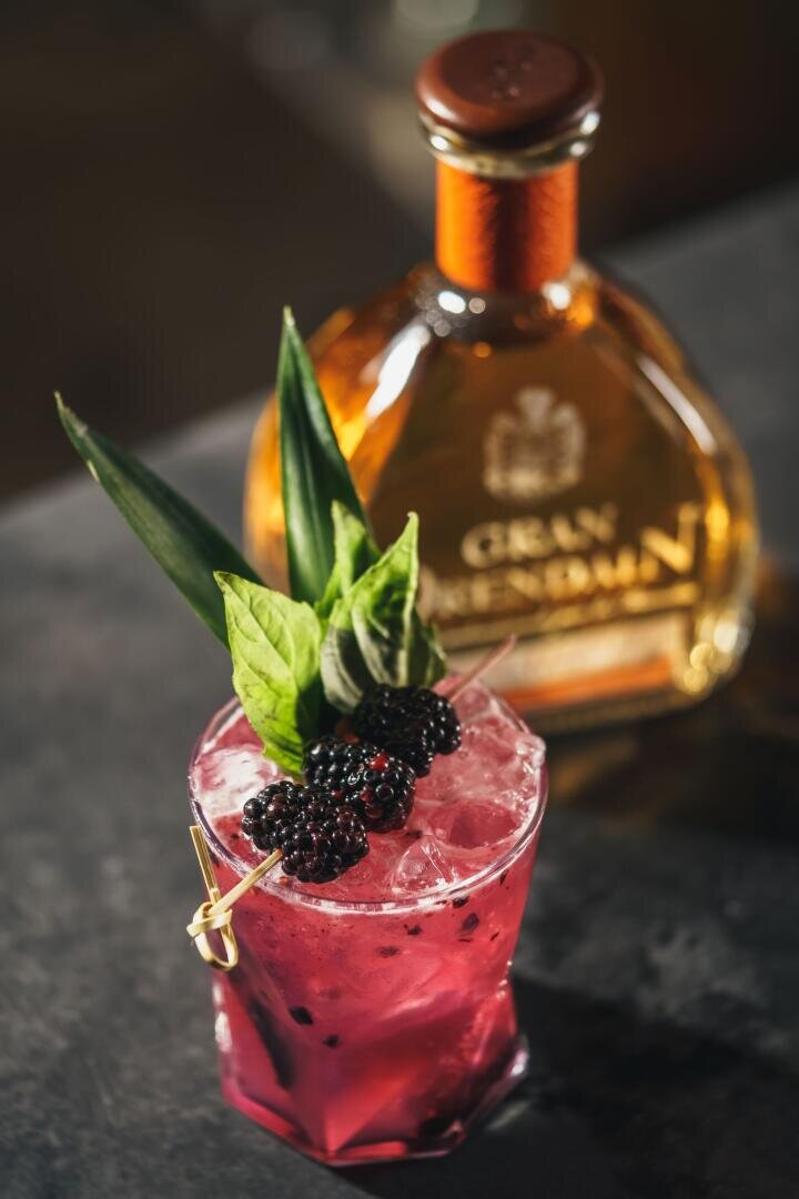 Ein leuchtend rosafarbener Cocktail mit Eis, garniert mit Brombeeren, Ananasblättern und frischen Kräutern, steht vor einer verschwommenen Flasche Gran Reserva auf einer dunklen Fläche.