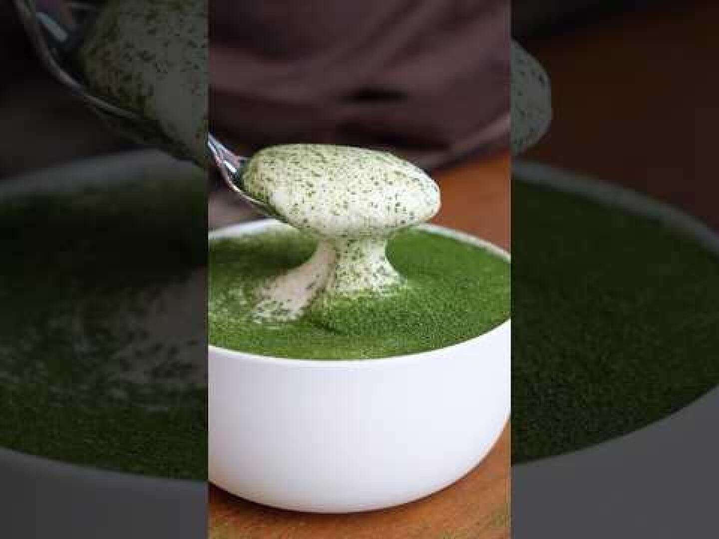 Eine weiße Schüssel mit einem cremigen Getränk, garniert mit grünem Matcha-Pulver. Ein Löffel hebt einen Klecks der schaumigen Mischung über die Schüssel und enthüllt so die dicke, glatte Konsistenz. Im Hintergrund ist eine braune Oberfläche zu sehen.
