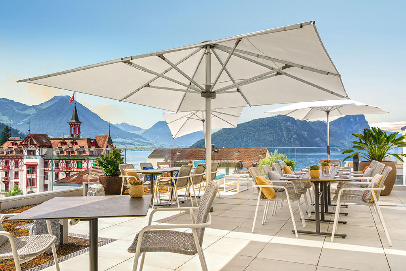 Eine sonnige Restaurantterrasse mit weißen Sonnenschirmen, modernen Tischen und Stühlen, mit Blick auf die Berge, einen See und ein historisches Gebäude mit roten Dächern im Hintergrund. Üppige Pflanzen begrünen den Sitzbereich.