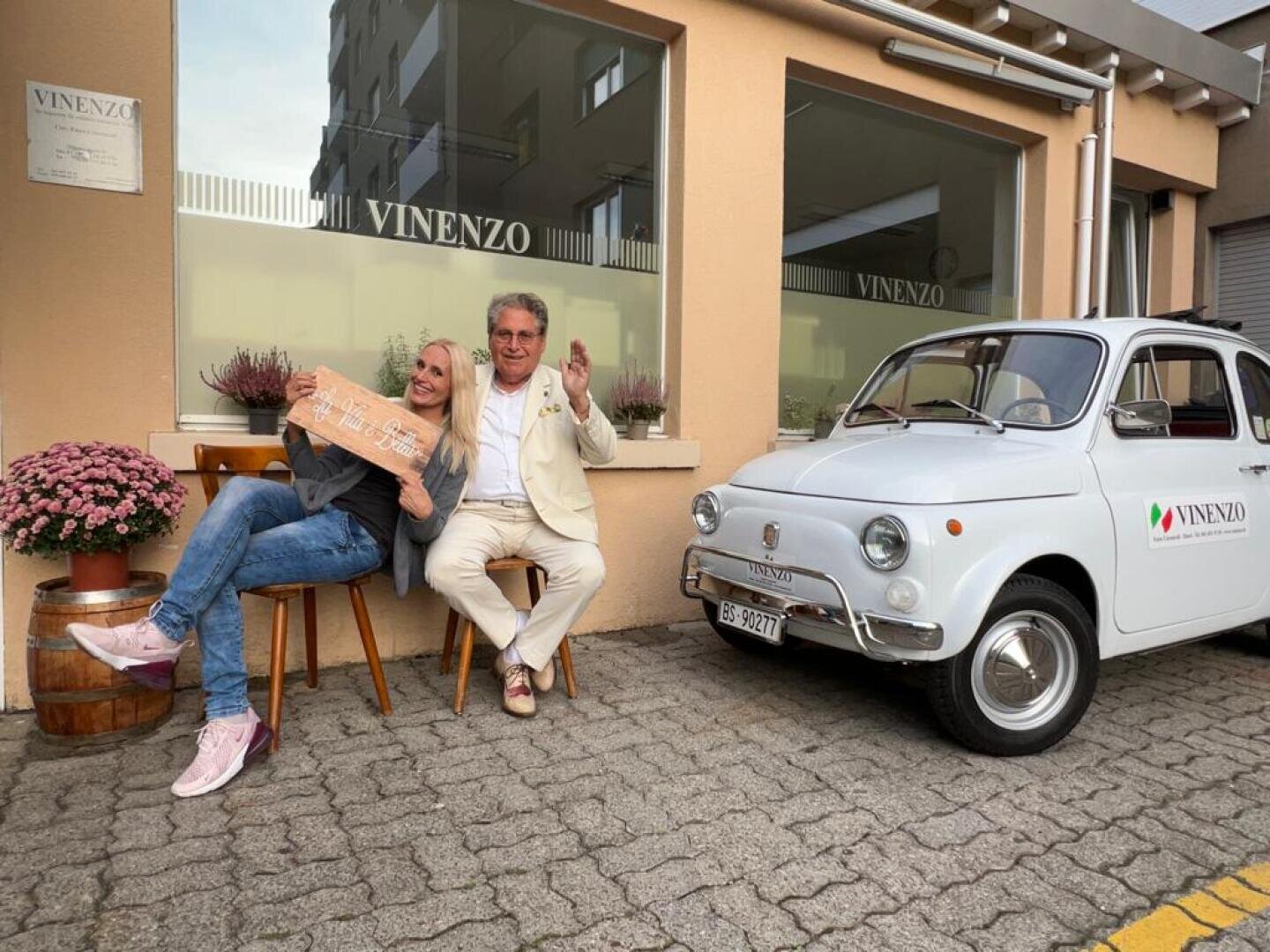 Zwei Personen lächeln und posieren auf einer Bank vor einem Gebäude mit der Aufschrift Vinenzo. Eine Person hält ein Holzschild in der Hand, während die andere ein Friedenszeichen macht. Ein weißer Oldtimer mit der Aufschrift Vinenzo ist in der Nähe geparkt. Blumen schmücken die Szene.