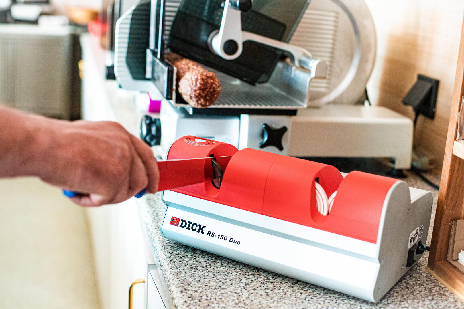 Eine Person schärft ein Messer mit einem rot-silbernen elektrischen Messerschärfer mit der Aufschrift DICK RS-150 Duo auf einem Küchentisch; im Hintergrund sind eine Aufschnittmaschine und Wurst zu sehen.