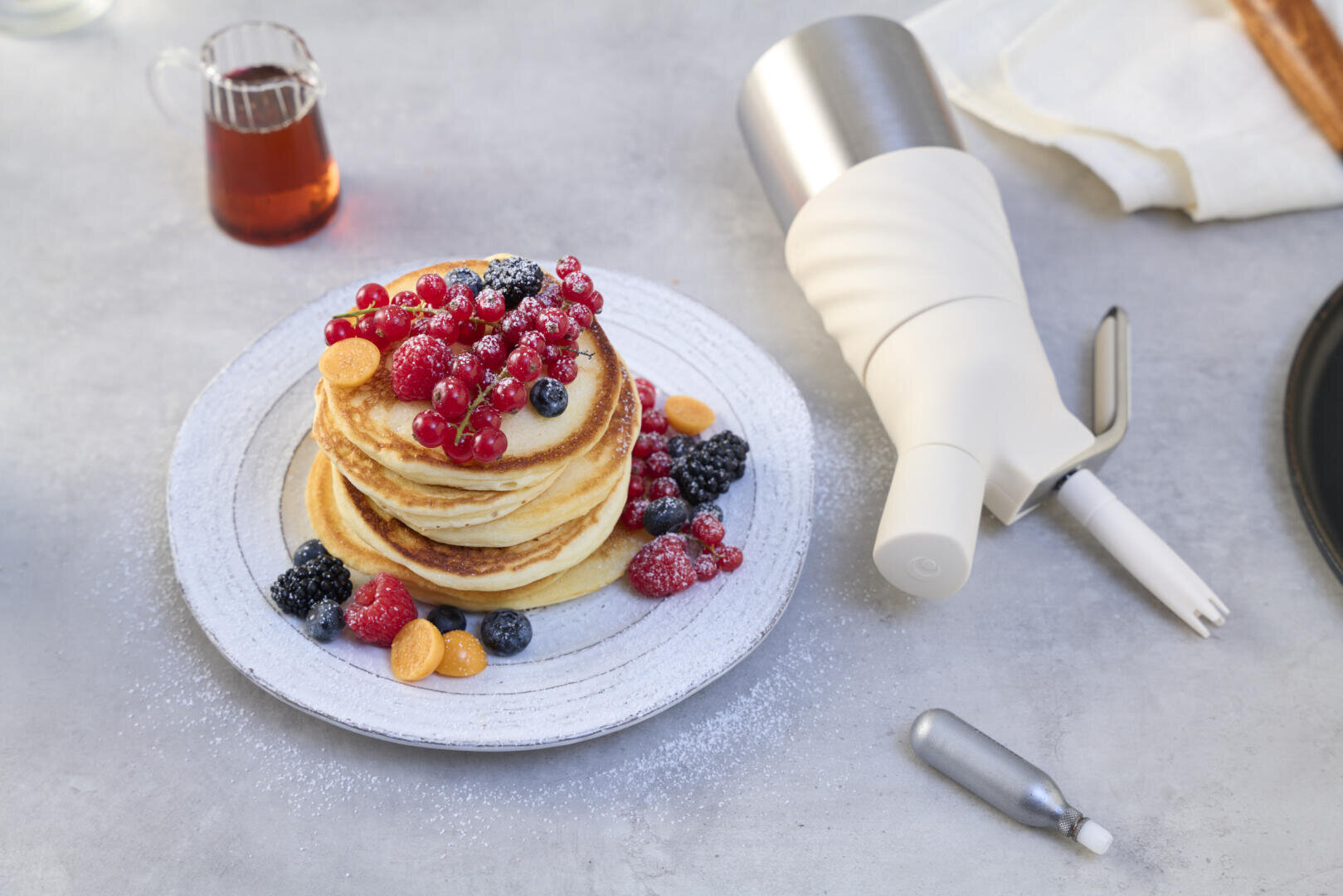 Ein Stapel Pfannkuchen mit verschiedenen Beeren und Puderzucker liegt auf einem Teller neben einem Sahnespender, einem kleinen Sirupkrug und einer Serviette auf einer hellgrauen Oberfläche.
